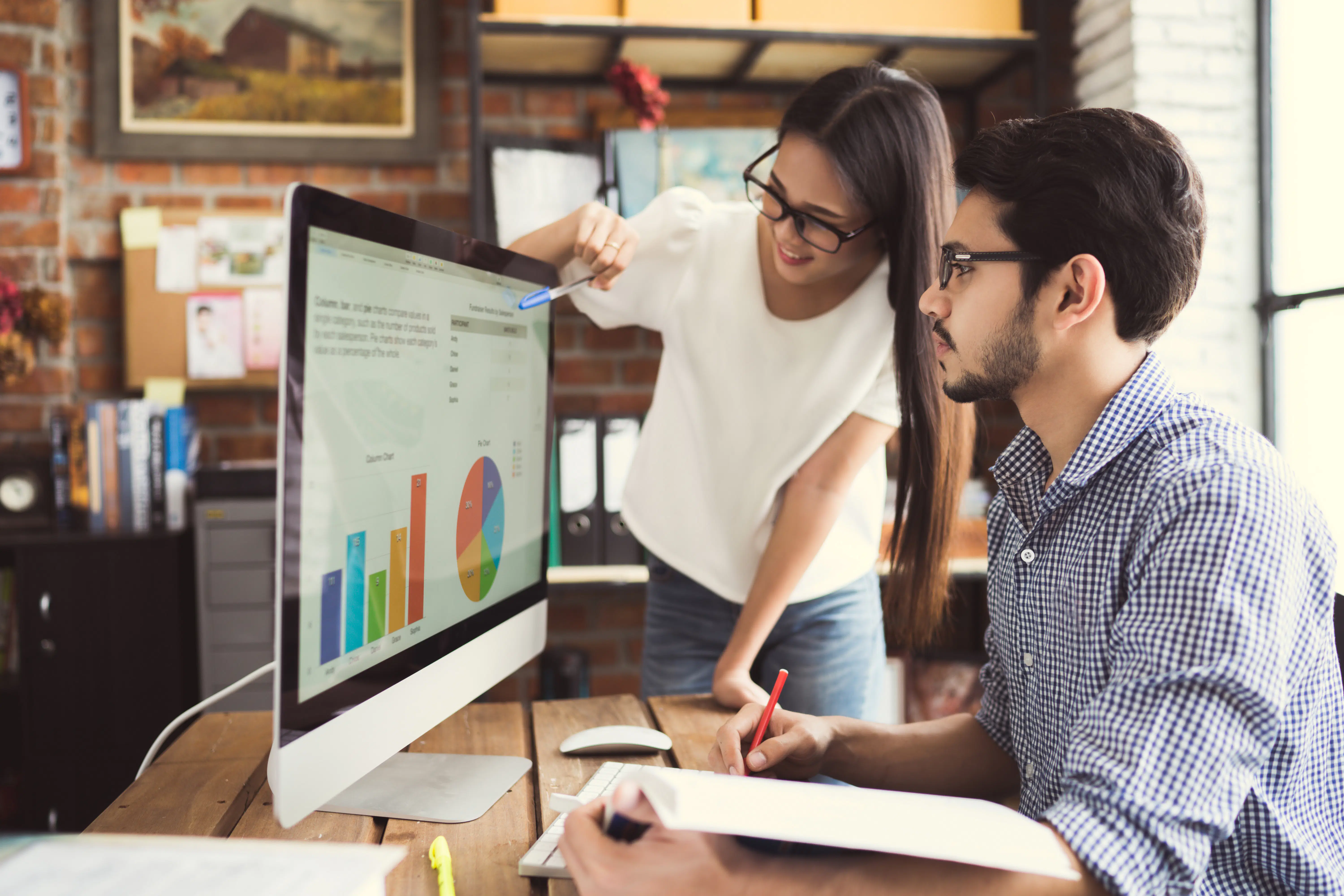 Two office colleagues discuss the charts results displayed on a computer screen